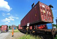 182_6946 Ein Container wird von einem Container Carrier auf den Gterwaggon eines Eisenbahnzugs geladen - ein Arbeiter in Signalkleidung und Helm berwacht die Arbeit. Auf der Strasse fhrt ein Sattelzug mit einem Containertransport.