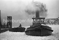 219_10_051 Ein Schlepper unter Dampf im Vulkanhafen - dick steigt der Qualm in den winterlichen Hamburger Himmel; das Wasser des Hamburger Hafens ist mit Treibeis bedeckt. Ein Schiffer auf einer Schute hat ein Tau in der Hand und bereitet sich darauf vor, das Schiff am Anleger festzumachen. Im Hintergrund Trmmer des U-Boot Bunkers Elbe 2; in dem 1941 fertig gestellten Bunker wurden die auf der Howaldtswerft gebauten U-Boote ausgerstet. Der Bunker hatte eine Deckenstrke von 3m - nach Kriegsende wurde die Anlage von britischen Truppen mit 47 Waggonladungen Fliegerbomben gesprengt.