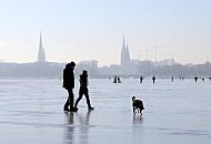 240_5501 Hamburger Spaziergnger nutzen den Sonnenschein und gehen mit ihrem Hund ber das blanke Eis der zugefrorenen Alster. Im Hintergrund die Kennedybrcke und die Trm der St. Petrikirchen und St. Nikolaikirche sowie der Turm des Hamburger Rathauses.