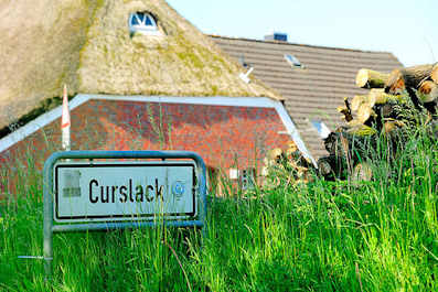 2835 Ortsschild CURSLACK - Holzstapel und Reetdach eines Bauernhauses.