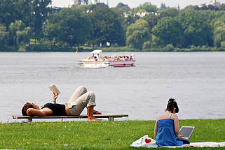 4b Entspannung am Ufer der Aussenalster - Ruhe vom Grossstadttrubel. Eine Frau liegt auf der Bank und liest ein Buch, eine andere sitzt auf der Wiese mit ihrem Labtop - im Hintergrund das Alstercabrio .