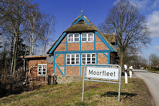 6716 Stadtteilschild am Strassenrand - reetgedecktes Fachwerkhaus mit blauen Balken.
