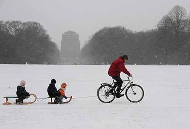 1174_5069 Winterzeit in der Hansestadt Hamburg - ein Vater zieht mit dem Fahrrad zwei Schlitten mit Kindern durch den Schnee im Stadtpark.