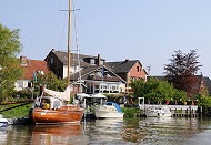 036_7450 Die Este ist ein reizvolles und interessantes Sportbootrevier. Viele Anwohner haben ihre Schiffe am Ufer liegen und fahren mit ihren Booten auf  der naheliegenden Elbe. Ein Holzsegelschiff und Sport-Motorboote liegen am Ufer der Este - im Hintergrund die Terrasse eines Cafs am Wasser.